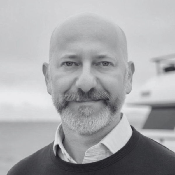 A man with a bald head and a beard smiles at the camera. Wearing a dark sweater over a collared shirt, he stands in front of water and part of a boat. This black and white image was taken for the Center for Architecture + Design.