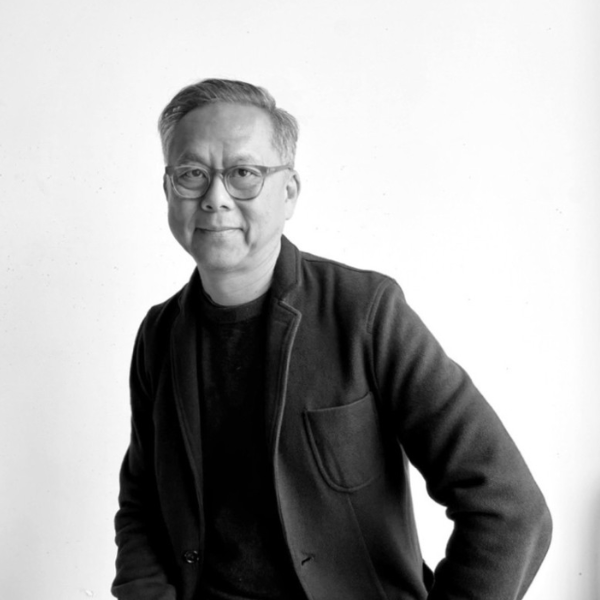 A man with short hair and glasses, wearing a dark jacket and sweater, sits against a plain white background, looking at the camera with a slight smile. The image is in black and white.
