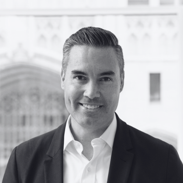 A man in a suit and white shirt smiles at the camera. The background, blurred, hints at large windows and architectural details of the Center for Architecture + Design. The photo is in black and white.