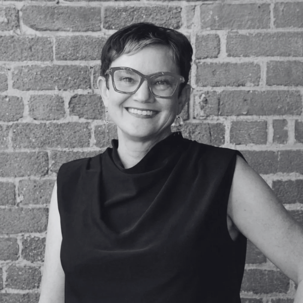 A woman with short dark hair and glasses, wearing a sleeveless black top, smiles while standing in front of a brick wall at the Center for Architecture + Design. The photo is in black and white.
