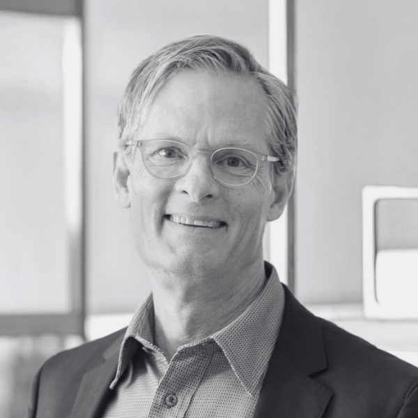 A man with light hair wearing glasses, a blazer, and a collared shirt smiles at the camera. The softly blurred indoor background hints at the Center for Architecture + Design. The image is in black and white.
