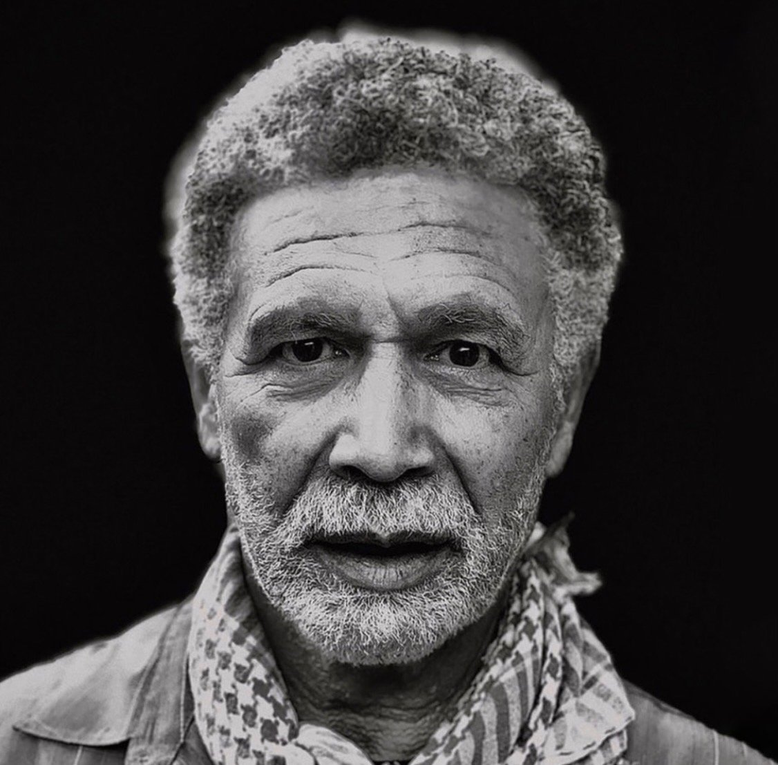 Black and white portrait of an older man with curly gray hair, a mustache, and beard. He is wearing a patterned scarf and a collared shirt, looking directly at the camera against a dark background.