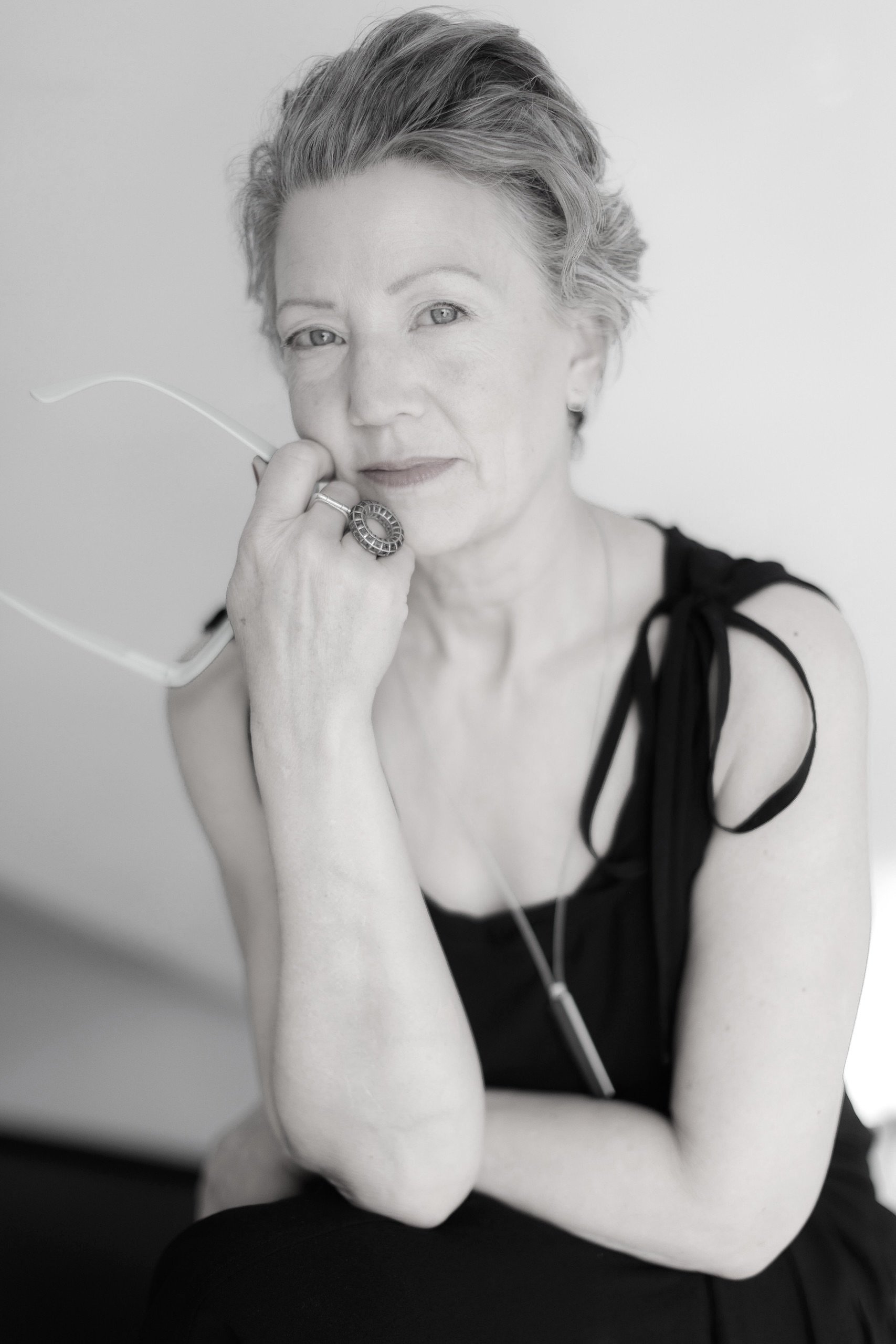 A woman with short, wavy gray hair wearing a sleeveless black outfit gazes at the camera, holding eyeglasses in one hand and resting her chin on her hand. The image is in black and white.