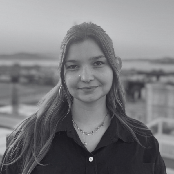 A young woman with long hair, wearing a dark shirt, a necklace, and hoop earrings, stands outdoors and smiles at the camera. The background is blurred, showing a cityscape and water in grayscale.