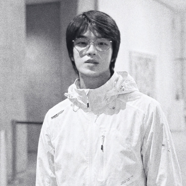 A young person with medium-length dark hair and glasses wears a light-colored jacket, standing indoors against a blurred background. The image is in black and white, giving it a grainy, vintage feel.