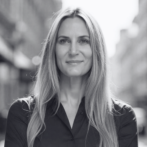 A woman with long, straight hair wearing a button-up shirt stands outdoors, smiling softly. The background is blurred, suggesting a city street near the Center for Architecture + Design, and the image is in black and white.