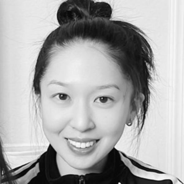 A woman with long dark hair tied in a bun is smiling at the camera. She is wearing a zip-up jacket and small earrings. The photo is in black and white.